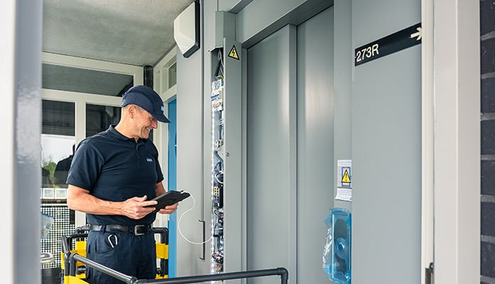Ein Aufzugstechniker mit blauem T-Shirt, Hose und Cap steht vor einem Aufzug und führt eine Wartung durch.