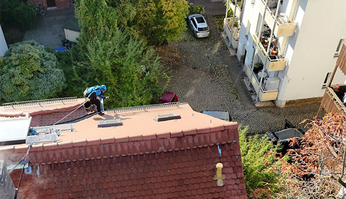 Blick von oben auf ein Hausdach mit roten Dachziegeln, auf dem ein Mitarbeiter, der mit einem Seil gesichert ist, die Dachrinne reinigt.