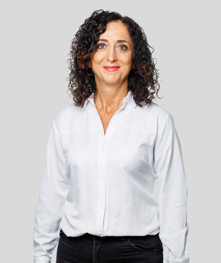 Portrait of a smiling woman with shoulder-length curly dark hair wearing a white button-down shirt against a light gray background.