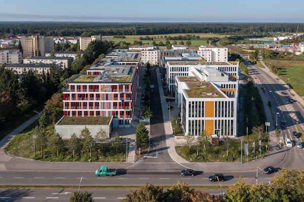 Aerial view of a modern office campus with two prominent white and red buildings, tree-lined avenues, and greenery on rooftops.