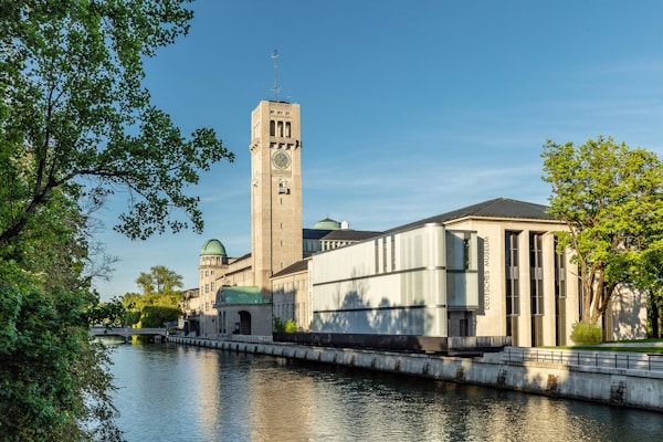 Riverside campus building with a tall clock tower beside a calm waterway and a modern glass wing, under a clear blue sky.