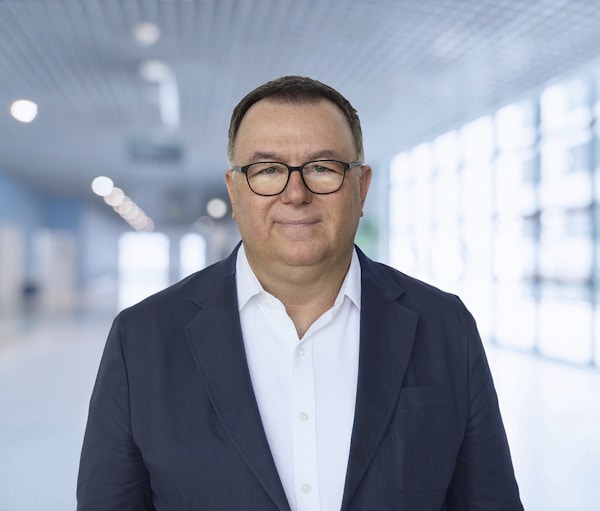 Portrait of a middle-aged man in a white shirt and navy blazer standing in a bright, modern hallway, looking at the camera.