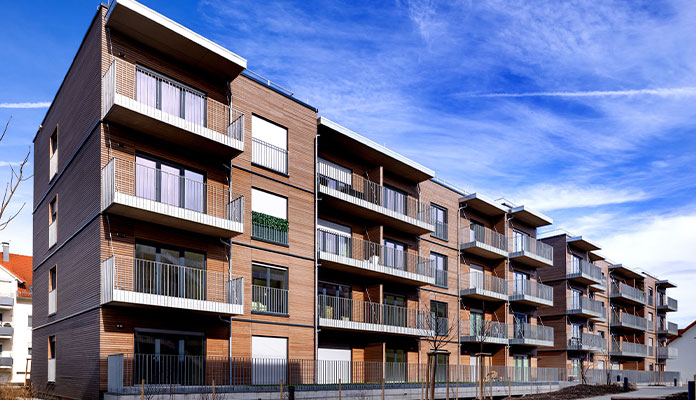 Das Bild zeigt ein modernes, mehrstöckiges Wohngebäude mit mehreren Balkonen. Die Fassade besteht hauptsächlich aus Holz und Glas, was dem Gebäude einen zeitgemäßen und freundlichen Eindruck verleiht. Das Gebäude hat drei Etagen und jede Wohnung verfügt über große Fenster und einen eigenen Balkon mit Metallgeländer. Vor dem Gebäude befinden sich junge, noch kleine Bäume und ein gepflasterter Bereich. Der Himmel ist blau mit einigen weißen Wolken und Kondensstreifen. Die Szene wirkt sauber und neu, typisch für einen modernen Wohnkomplex in einer Stadt oder Vorstadt.