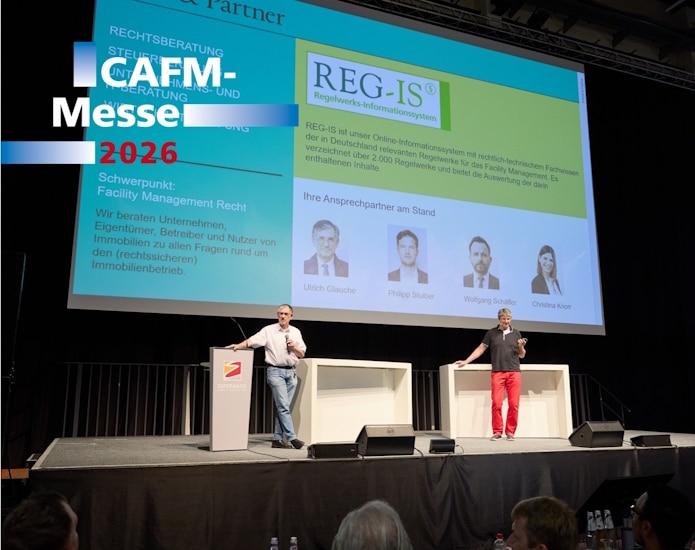 Two men present on a stage at a conference with a large screen showing a REG-IS information system and speaker photos.