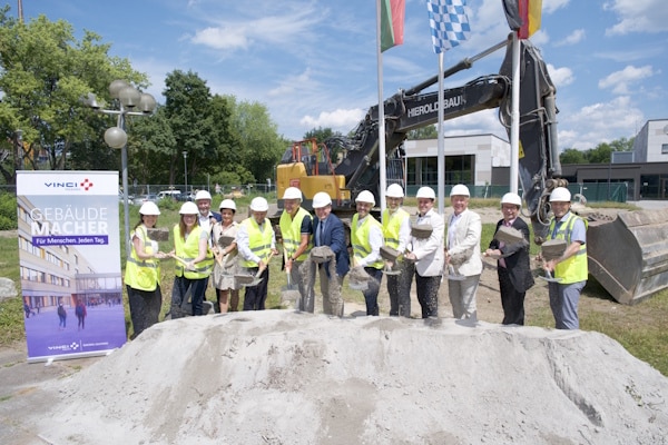 Group of people in hard hats and safety vests at a construction site, posing behind a mound of sand with a large excavator in the background.