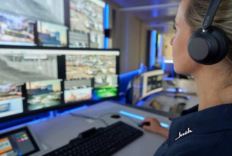 Operator wearing headphones at a multi-monitor surveillance station, monitoring video feeds. Requirements: concise, informative, and function-focused.
