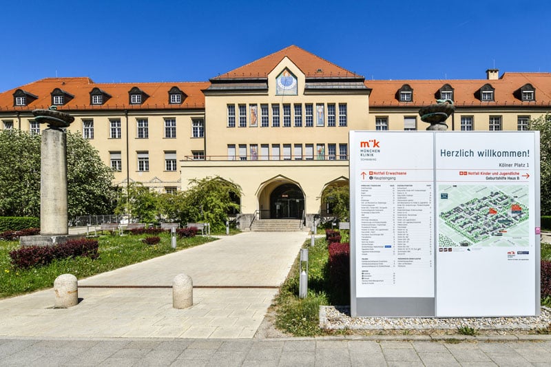 Beige campus building with a red roof and arched entrance; a large welcome/information signboard with a map stands to the right.