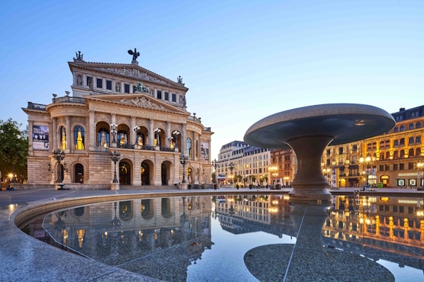 Grand neoclassical building with columns and statues, reflected in a circular fountain in a city square at dusk.
