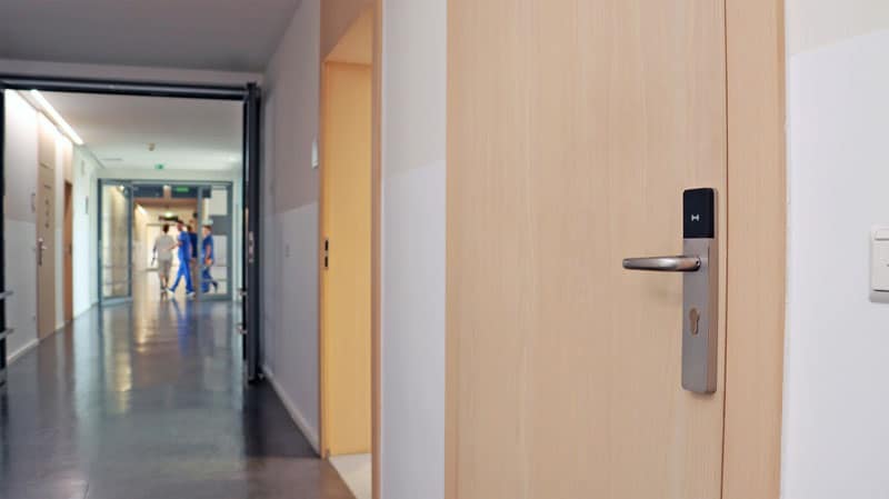 Hospital hallway with a wooden door on the right and staff in blue scrubs walking in the distance