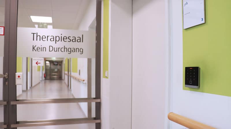 Glass door with a 'Therapiesaal Kein Durchgang' sign; an empty hospital corridor extends beyond it into the distance.