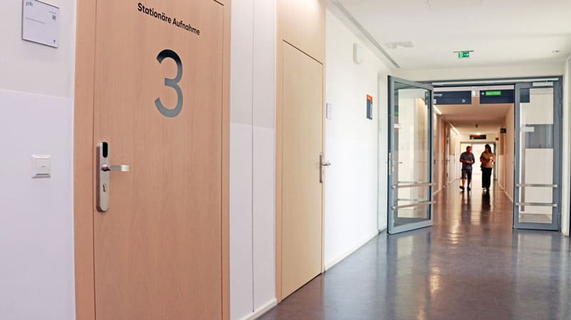 Hospital corridor with a wooden door labeled '3' and 'Stationäre Aufnahme', glass doors at the far end, and two people walking in the distance.