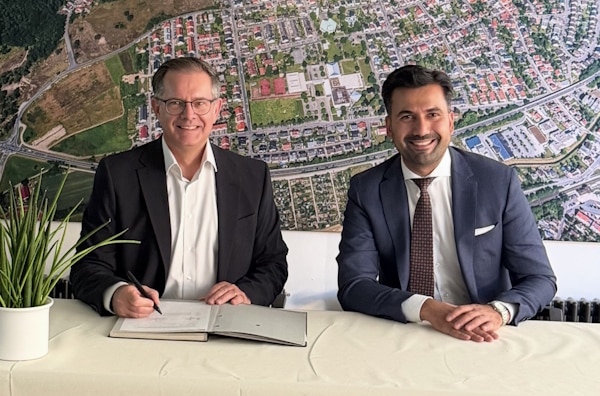 Two men sit at a table signing a document, with a large aerial map on the wall behind them.