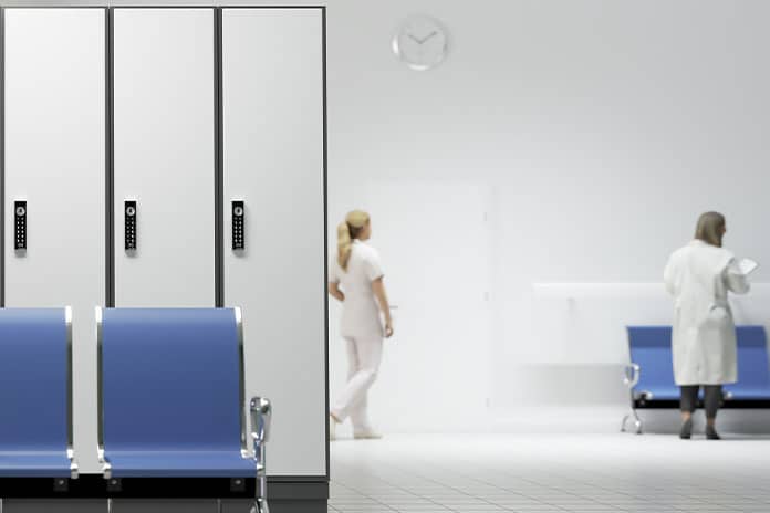 Hospital hallway with three tall grey lockers on the left and blue waiting chairs; a nurse and a doctor appear blurred in the background.