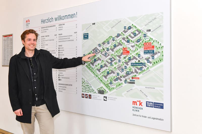 Man in a black coat points to a large hospital map and directory on a wall in a medical facility (German text reads 'Herzlich willkommen!').