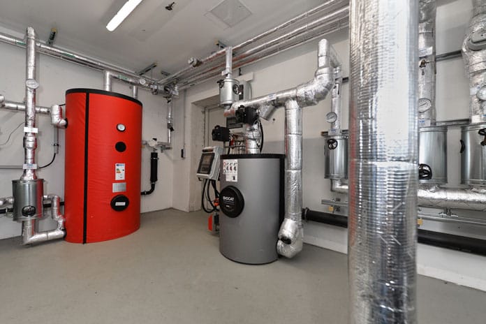 Industrial boiler room with a tall red hot-water tank and a gray boiler, surrounded by insulated metal pipes and controls at ceiling level.