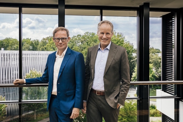 Two business colleagues in suits stand by a glass railing in a modern office atrium, smiling at the camera, with greenery outside.