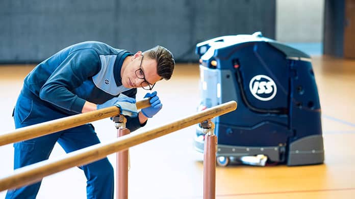 ISS-Mitarbeiter reinigt einen Barren in der Turnhalle, während ein autonomer Reinigungsroboter im Hintergrund arbeitet.