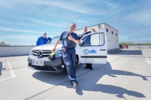 Three smiling workers in blue uniforms pose with a white SFC Group van on a sunny rooftop parking lot.