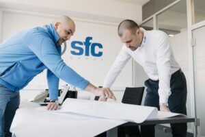 Two men lean over a table, examining a large blueprint together in a bright office.