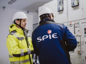 Two workers in hard hats and high-visibility jackets inspect a large electrical control panel.